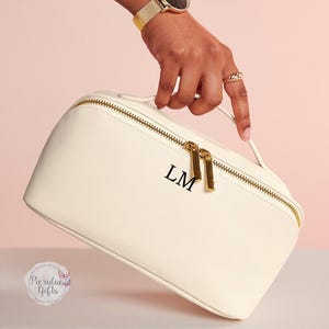 May include: Cream-colored cosmetic bag with gold-tone zipper and handle. The bag features the initials "LM" in black. The bag is held by a person wearing a gold watch and ring. The bag is on a light pink and white background.