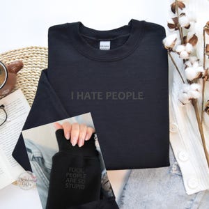 May include: Black crew neck sweatshirt with the text "I HATE PEOPLE" printed on the front. The sleeve cuff displays the words "FUCK, PEOPLE ARE SO STUPID". The sweatshirt is folded, with a coffee cup and book in the background.