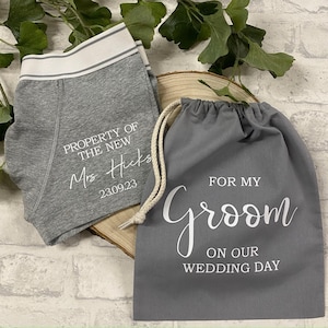 May include: Gray boxer briefs with white trim and text reading "PROPERTY OF THE NEW Mrs. Hicks 23.09.23" next to a gray drawstring bag with "FOR MY Groom ON OUR WEDDING DAY" in white.