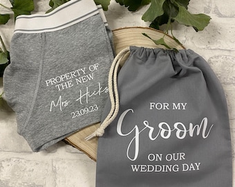 May include: Grey boxer briefs with a white waistband and the text PROPERTY OF THE NEW Mrs. Hicks 23.09.23, paired with a dark grey drawstring fabric bag featuring the white text FOR MY groom ON OUR WEDDING DAY printed on the front surface.