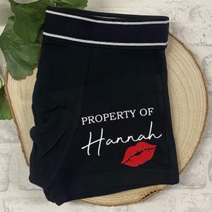 May include: Black boxer shorts with a white and silver striped waistband. The text "PROPERTY OF Hannah" is printed in white, with a red lip graphic below. The shorts are displayed on a wooden slice.