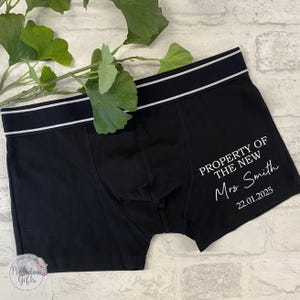 May include: Black boxer briefs featuring a white elastic waistband with two thin horizontal stripes. The right side displays white text reading PROPERTY OF THE NEW Mrs Smith followed by the date 22.01.2025 printed in a clean, sans-serif and script font.