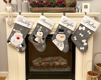 May include: Four gray Christmas stockings with white trim and embroidered names: Daddy, Mummy, Leyla, and Pedro. Each stocking features a different festive character: Santa Claus, a penguin, a snowman, and a bone.
