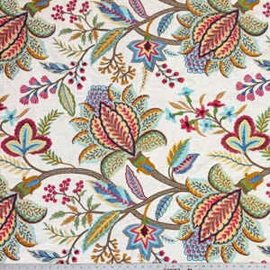 May include: A white fabric featuring a repeating floral pattern with large stylized blossoms in shades of magenta, teal, yellow, and brown. The design includes intricate leafy vines and small berry clusters, with a visible ruler at the bottom edge.