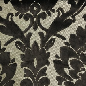 Pu&ograve; includere: Un tessuto caratterizzato da un motivo damascato in velluto nero su sfondo beige chiaro. Il materiale presenta un motivo floreale simmetrico ripetuto con texture morbide e in rilievo che contrastano con la base liscia e intrecciata.