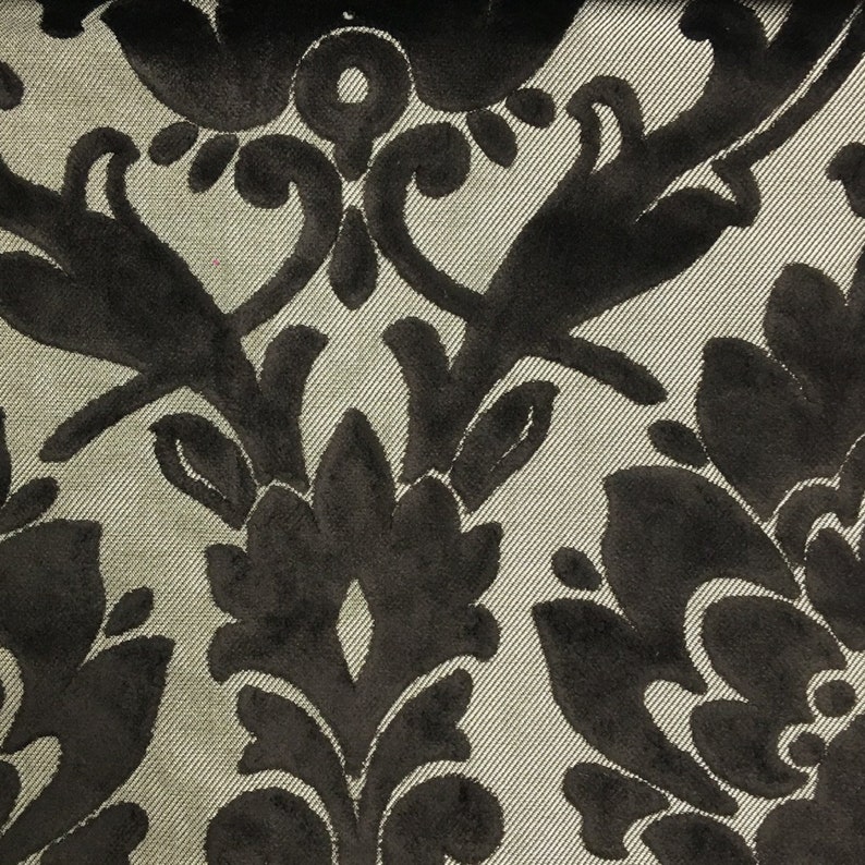 Pu&ograve; includere: Un tessuto caratterizzato da un motivo damascato in velluto nero su sfondo beige chiaro. Il materiale presenta un motivo floreale simmetrico ripetuto con texture morbide e in rilievo che contrastano con la base liscia e intrecciata.