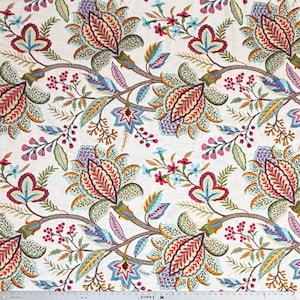 May include: A white fabric featuring a repeating floral pattern with large stylized blossoms in shades of red, blue, yellow, and green. A metal ruler is visible at the bottom edge, showing measurements in inches for scale and reference.