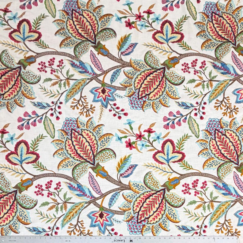 May include: A white fabric featuring a repeating floral pattern with large stylized blossoms in shades of red, blue, yellow, and green. A metal ruler is visible at the bottom edge, showing measurements in inches for scale and reference.