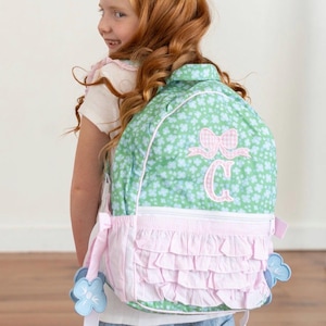 May include: A light green backpack featuring a white clover pattern, a pink gingham bow, and a pink letter C monogram. The bag includes a white zippered front pocket with layered pink ruffles and light blue flower-shaped zipper pulls.