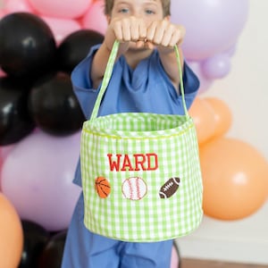 Op de afbeelding: Een groen-witte geruite stoffen draagtas met de naam "WARD" in het rood geborduurd. De tas heeft geborduurde sportbalontwerpen: een basketbal, een honkbal en een voetbal. De tas wordt vastgehouden door een persoon in een blauw shirt.