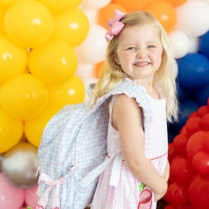 Peut inclure: Une jeune fille porte une robe rose &agrave; carreaux vichy et un sac &agrave; dos assorti. Le sac &agrave; dos est bleu clair avec un motif floral et des n&oelig;uds roses. En arri&egrave;re-plan, un mur de ballons color&eacute;s jaunes, oranges, blancs, bleus et rouges.