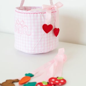 May include: A pink and white gingham Easter basket with a matching handle and bow. The basket has a scalloped edge and red heart accents. Attached are felt food shapes including carrots, apples, and trees.