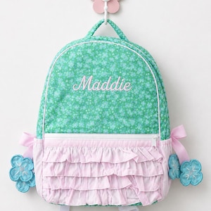 May include: A green backpack with a small floral pattern and the name Maddie embroidered in pink. The front features a white zippered pocket with three horizontal rows of light pink ruffled fabric. Two blue flower appliqués hang from the sides.