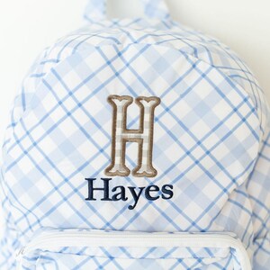 Peut inclure: Sac &agrave; dos &agrave; carreaux bleus et blancs avec une poche zipp&eacute;e blanche. Il pr&eacute;sente une lettre "H" brod&eacute;e en marron et le nom "Hayes" en bleu marine.