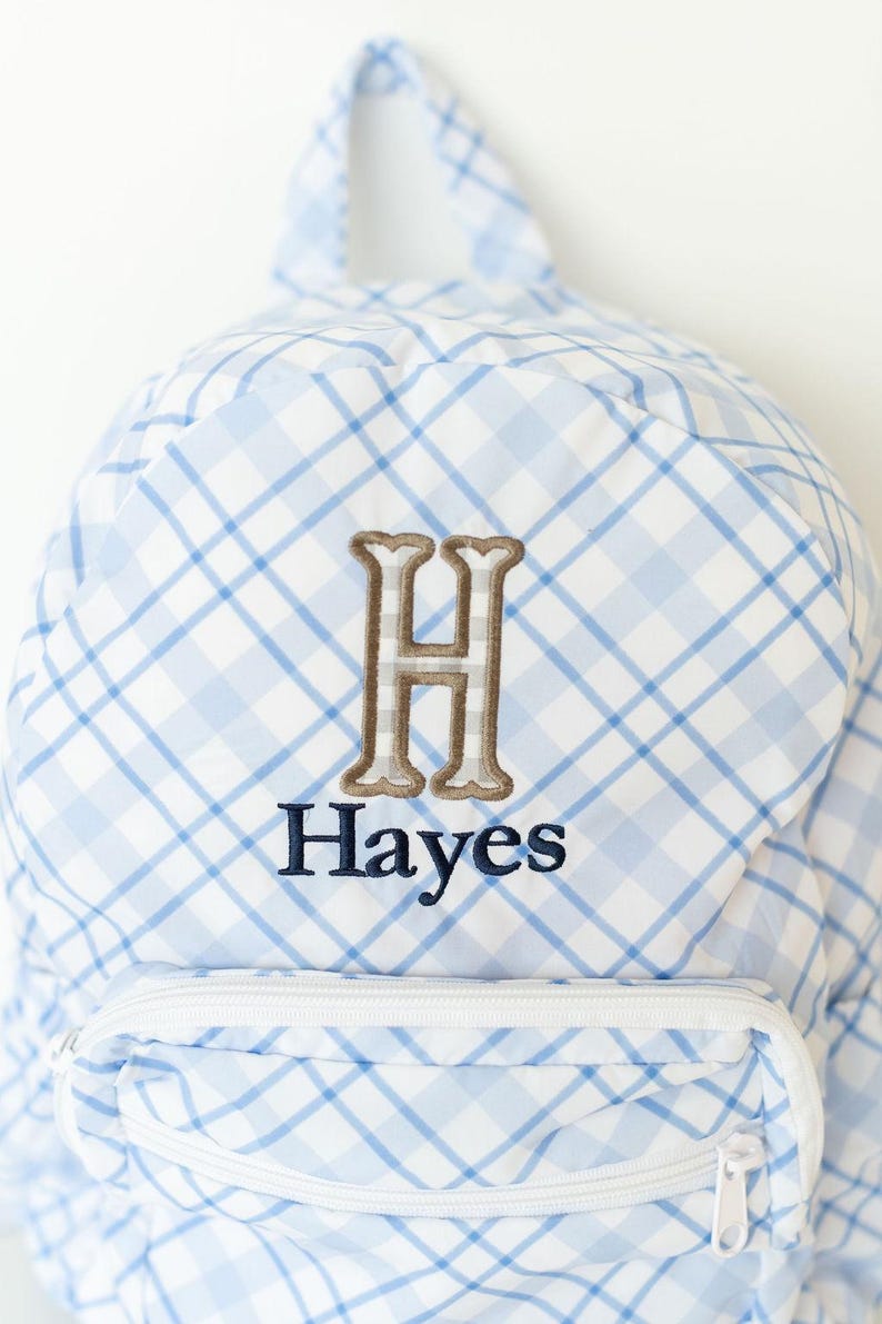 Peut inclure: Sac &agrave; dos &agrave; carreaux bleus et blancs avec une poche zipp&eacute;e blanche. Il pr&eacute;sente une lettre "H" brod&eacute;e en marron et le nom "Hayes" en bleu marine.