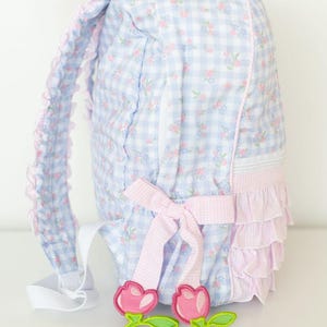 Peut inclure: Un sac &agrave; dos &agrave; carreaux bleus et blancs avec des d&eacute;tails floraux roses et un n&oelig;ud rose. Le sac &agrave; dos a une bordure &agrave; volants et des appliques de fleurs roses d&eacute;coratives. Les bretelles sont blanches.