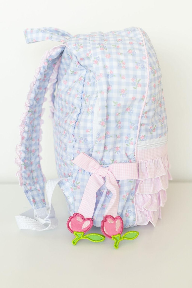 Peut inclure: Un sac &agrave; dos &agrave; carreaux bleus et blancs avec des d&eacute;tails floraux roses et un n&oelig;ud rose. Le sac &agrave; dos a une bordure &agrave; volants et des appliques de fleurs roses d&eacute;coratives. Les bretelles sont blanches.