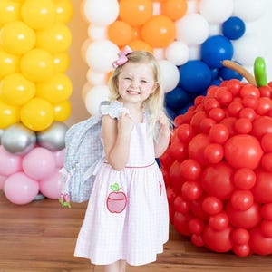 Peut inclure: Une jeune enfant porte une robe rose &agrave; carreaux avec une application de pomme et un sac &agrave; dos bleu clair. En arri&egrave;re-plan, des compositions de ballons color&eacute;s jaunes, oranges, blancs, bleus et roses, et une grande sculpture de pomme rouge.
