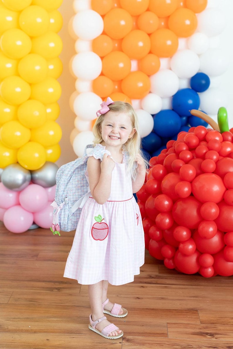 Peut inclure: Une jeune enfant porte une robe rose &agrave; carreaux avec une application de pomme et un sac &agrave; dos bleu clair. En arri&egrave;re-plan, des compositions de ballons color&eacute;s jaunes, oranges, blancs, bleus et roses, et une grande sculpture de pomme rouge.