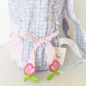 Peut inclure: Un sac &agrave; dos &agrave; carreaux bleu clair et blanc avec un motif floral. Le sac &agrave; dos a des accents roses, un n&oelig;ud et une sangle blanche. Deux appliqu&eacute;s en forme de tulipe rose avec des tiges vertes sont en bas. Une &eacute;tiquette avec le texte "Sweet Boutique" est attach&eacute;e.