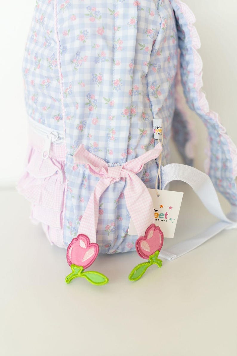 Peut inclure: Un sac &agrave; dos &agrave; carreaux bleu clair et blanc avec un motif floral. Le sac &agrave; dos a des accents roses, un n&oelig;ud et une sangle blanche. Deux appliqu&eacute;s en forme de tulipe rose avec des tiges vertes sont en bas. Une &eacute;tiquette avec le texte "Sweet Boutique" est attach&eacute;e.