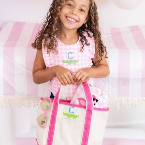 May include: A young girl holds a pink and white canvas tote bag with a matching shirt. The tote bag has a white body, pink trim, and a green alligator design with the letter "C". The shirt is pink gingham with a green alligator and the letter "C".