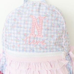 May include: A light blue and white gingham backpack with a floral pattern and pink accents. The backpack features a pink embroidered letter "N" and the name "Nere". The bottom has a pink ruffle design.