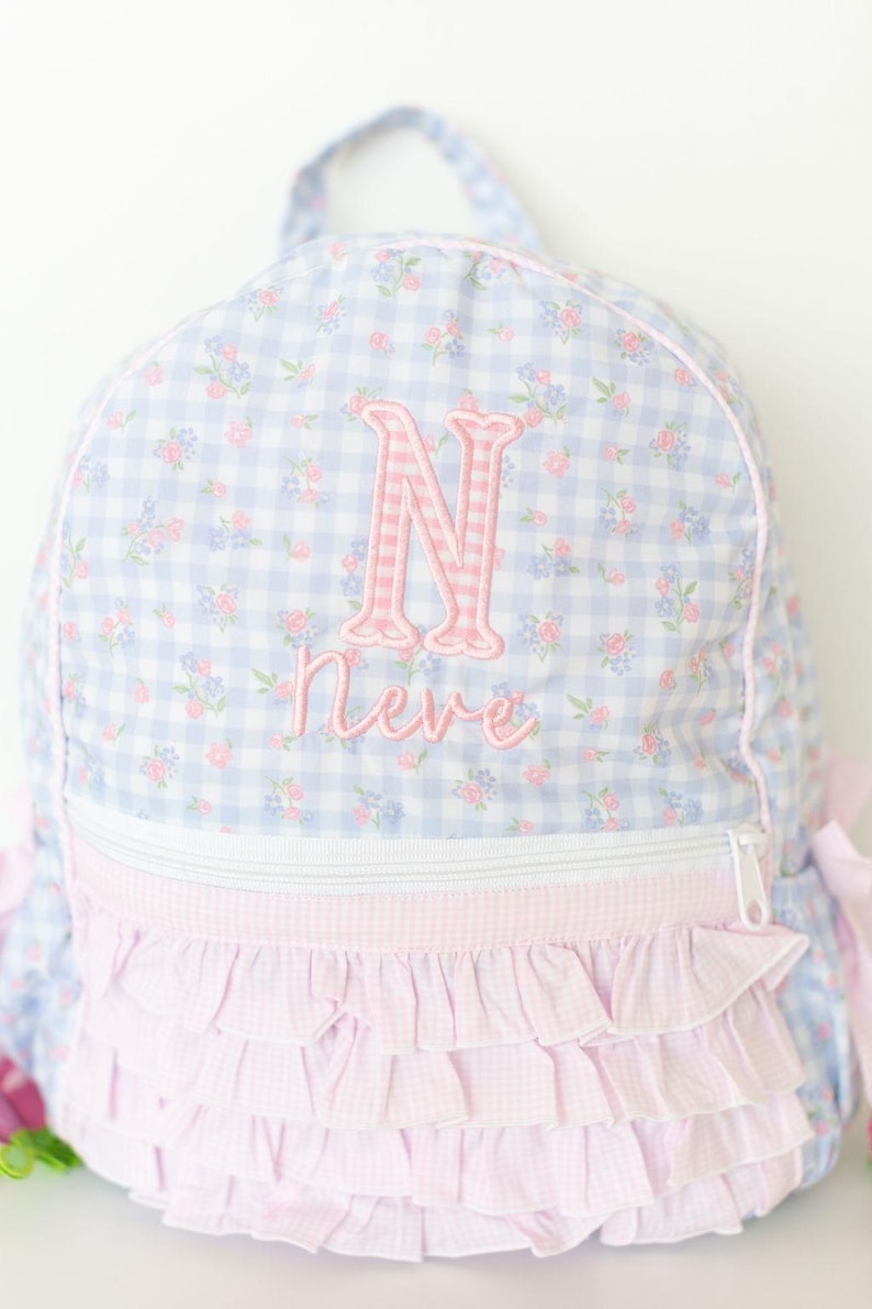 Peut inclure: Un sac &agrave; dos &agrave; carreaux bleu clair et blanc avec un motif floral et des accents roses. Le sac &agrave; dos pr&eacute;sente une lettre "N" rose brod&eacute;e et le nom "Neve". Le bas a un design &agrave; volants roses.