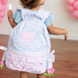 May include: A light blue and white gingham backpack with a pink heart and the name "Naomi" embroidered on it. The backpack has pink ruffles and flower accents. The child is wearing a matching dress.