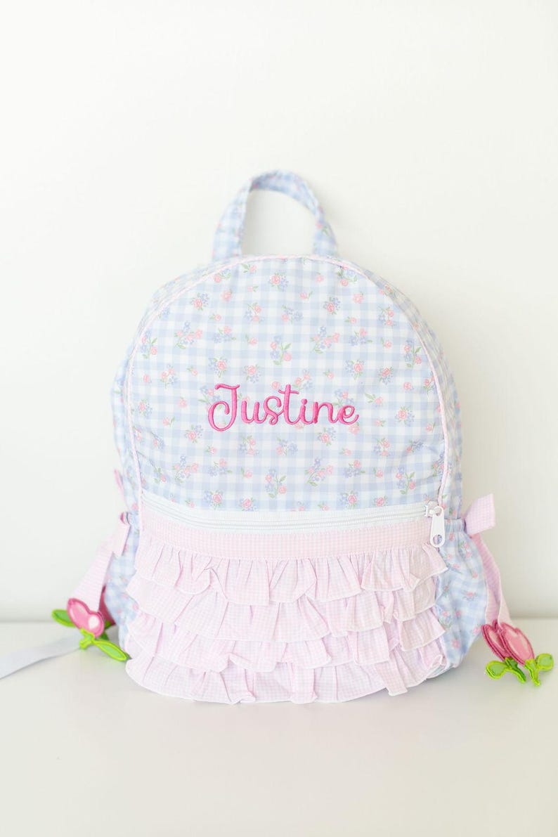 Peut inclure: Un sac &agrave; dos &agrave; carreaux bleus et blancs avec des motifs floraux et des volants roses. Le nom "Justine" est brod&eacute; en rose. Il comporte une poign&eacute;e sup&eacute;rieure, une fermeture &eacute;clair et des d&eacute;corations florales.