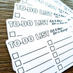 May include: A stack of white To-Do List notepads with black text. Each notepad has the words "TO-DO LIST" at the top, followed by lines and check boxes. Some notepads have the phrase "Get it done like a champ!" written in cursive.