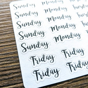 May include: A sheet of clear stickers with the days of the week written in black cursive lettering. The stickers are arranged in rows and columns, with each day of the week repeated multiple times.