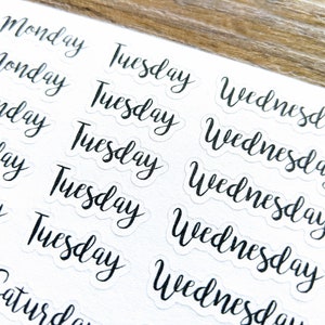 May include: A sheet of white paper with black handwritten stickers of the days of the week. The stickers say "Monday", "Tuesday", "Wednesday", "Thursday", "Friday", "Saturday", and "Sunday".
