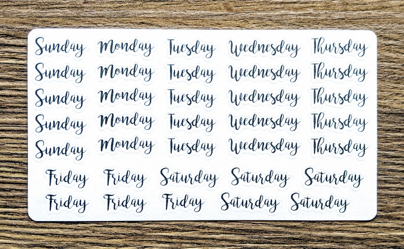 May include: A sheet of white stickers with black text. The stickers feature the days of the week, including Sunday, Monday, Tuesday, Wednesday, Thursday, Friday, and Saturday. Each day of the week is repeated five times on the sheet.