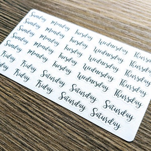 May include: A sheet of white stickers with black lettering. The stickers feature the days of the week, Sunday through Saturday, in a handwritten font.
