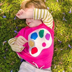 May include: A child's bright pink long-sleeved shirt with a white artist's palette design featuring colourful paint splotches. The sleeves have yellow and white stripes. The child is lying on green grass.