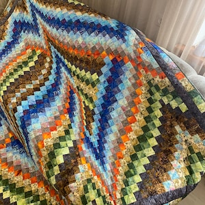May include: A handmade quilt with a vibrant, geometric pattern. The quilt features a patchwork design with squares in shades of blue, green, orange, and brown. The quilt is draped over a sofa, showcasing its texture and color variations.