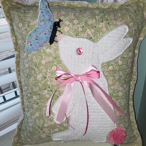 May include: A square decorative pillow with a white bunny applique and a light blue butterfly. The pillow has a green floral pattern and a pink ribbon bow. The bunny has a pink button eye and a pink flower accent.