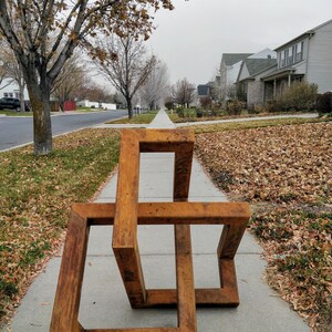 May include: A rusty brown metal sculpture of a three-dimensional geometric shape, resembling a cube or a box, with multiple intersecting planes. The sculpture is placed on a sidewalk in front of a residential neighborhood.