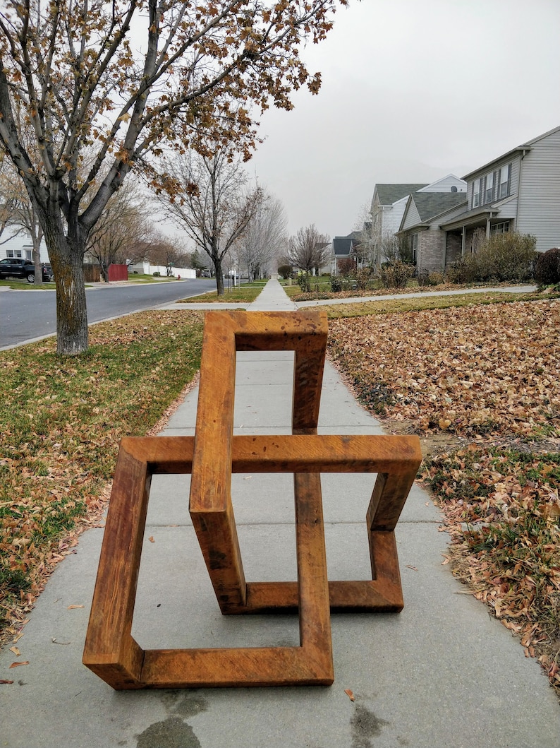 May include: A rusty brown metal sculpture of a three-dimensional geometric shape, resembling a cube or a box, with multiple intersecting planes. The sculpture is placed on a sidewalk in front of a residential neighborhood.