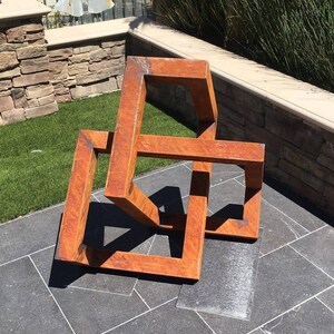 May include: A rusty metal sculpture of a three-dimensional geometric shape, resembling a cube or a box, with multiple intersecting planes. The sculpture is placed on a gray tiled surface.