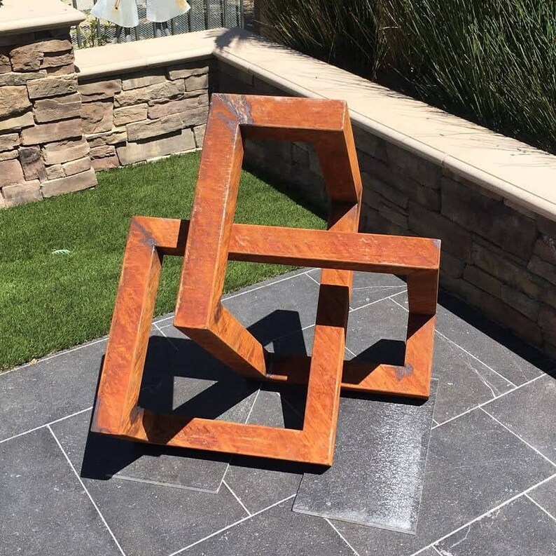 May include: A rusty metal sculpture of a three-dimensional geometric shape, resembling a cube or a box, with multiple intersecting planes. The sculpture is placed on a gray tiled surface.