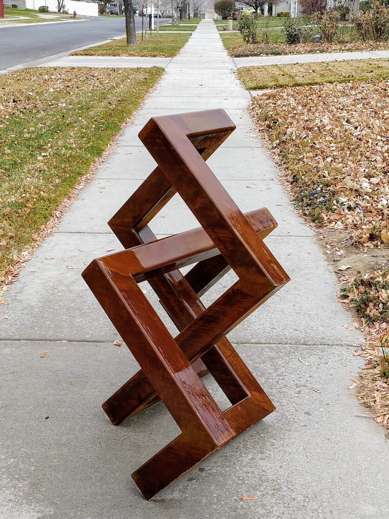 May include: A wooden sculpture made of four connected squares, each with a different orientation. The sculpture is a dark brown color and has a glossy finish. It is sitting on a concrete sidewalk.