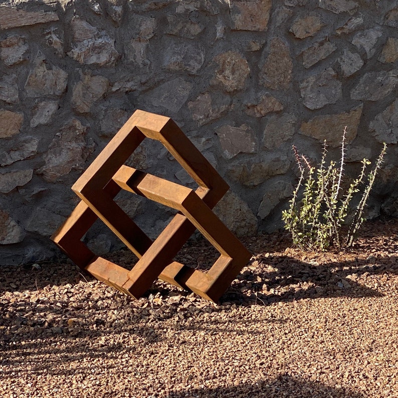 May include: A rusty metal sculpture of a geometric shape, resembling a cube or a diamond, with a three-dimensional design. The sculpture is placed on a bed of small brown rocks.