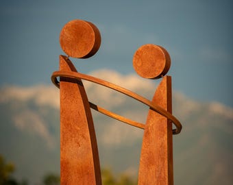 May include: A weathered metal sculpture featuring two vertical, rectangular forms topped with circular elements. A circular band connects the forms. The sculpture is set against a backdrop of a mountain range and a blue sky.