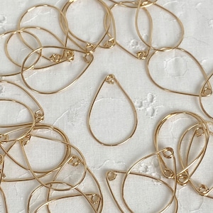 May include: A group of gold teardrop-shaped wire earring hoops. The hoops are arranged in a random pattern on a white fabric background.