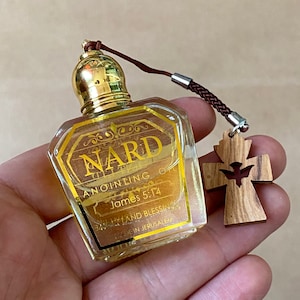 May include: A small, clear glass bottle of Nard Anointing Oil with a gold cap and label. The label includes text and decorative elements. A wooden cross pendant with a dove cutout hangs from a brown cord.