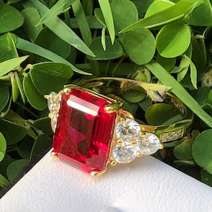 Może przedstawiać: Złoty pierścionek z dużym, czerwonym, szmaragdowo oszlifowanym kamieniem szlachetnym otoczonym mniejszymi, przezroczystymi kamieniami szlachetnymi. Pierścionek leży na białym tle.