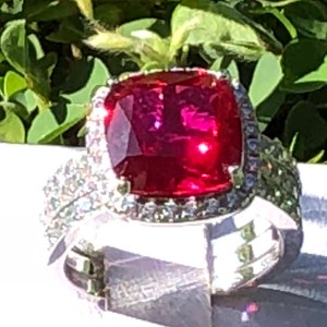 Puede incluir: Un anillo con una gran piedra preciosa roja de talla cojín, rodeada por un halo de piedras preciosas más pequeñas y transparentes. El anillo está engastado en plata.