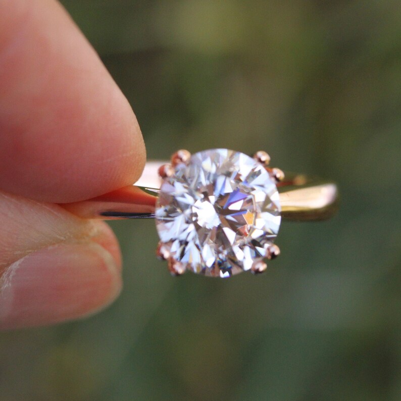 May include: A close-up of a rose gold engagement ring with a large, round, clear diamond.